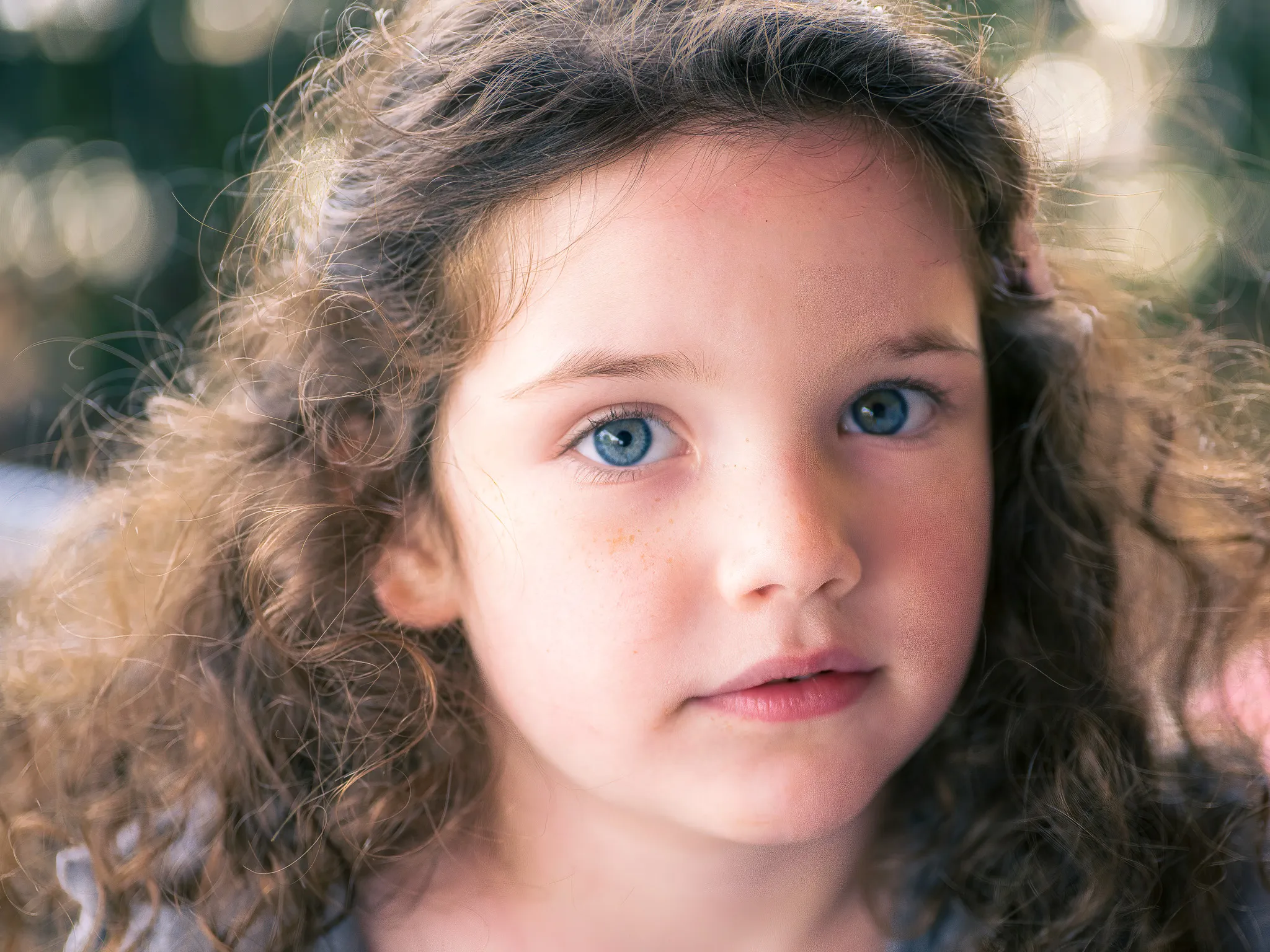 Close-up outdoor portrait of a young girl with curly brown hair and bright blue eyes in golden bokeh light