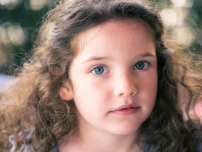 Close-up outdoor portrait of a young girl with curly brown hair and bright blue eyes in golden bokeh light