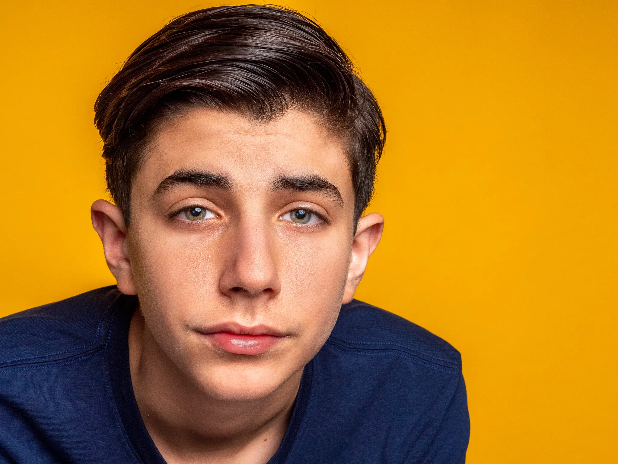 Teen boy with dark hair and blue eyes in navy shirt against bright yellow studio backdrop