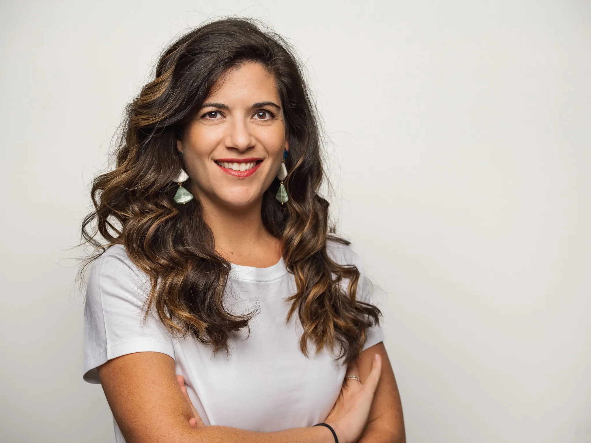Woman in her thirties with long wavy brown hair and statement earrings smiling with arms crossed against a light gray studio backdrop