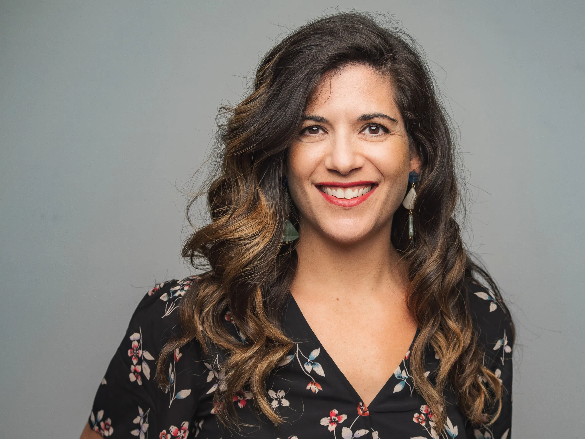 Woman in her thirties with long wavy brown hair wearing a black floral blouse and tassel earrings smiling against a gray studio backdrop