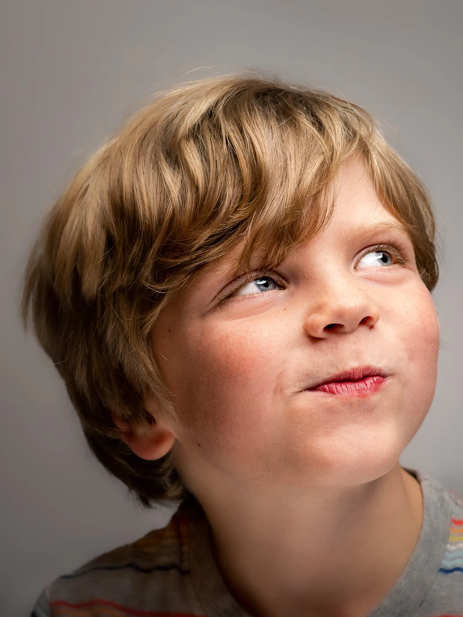 Close-up studio portrait of a young boy around age 5 with blonde hair and blue eyes looking upward, gray background