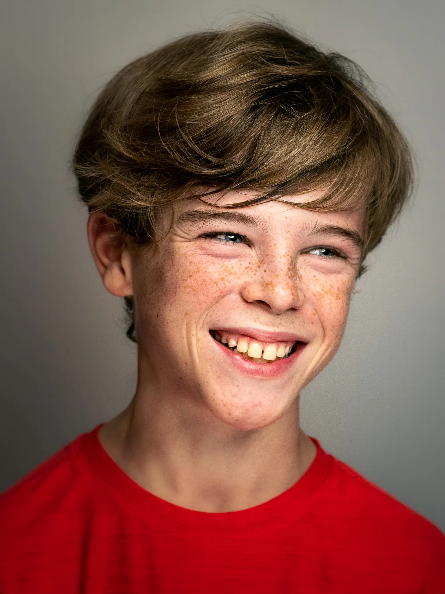 Freckled teenage boy in a red shirt smiling broadly in a studio portrait against a gray background