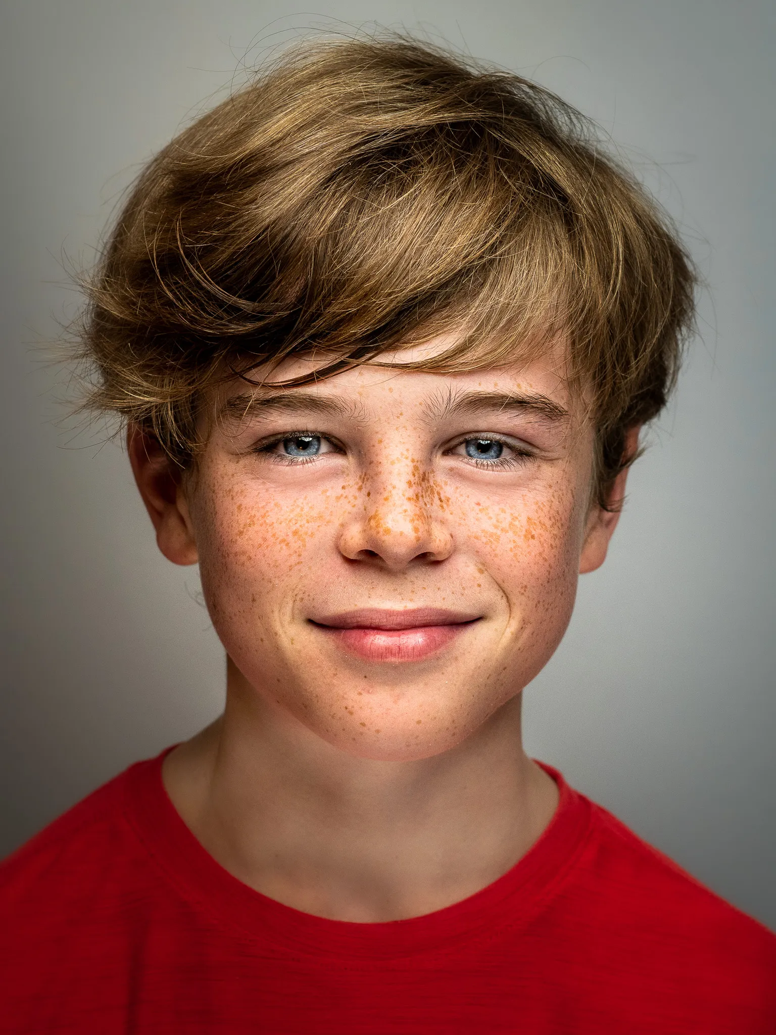Freckled boy around 12 years old with sandy blond hair and blue eyes smiling in a red shirt, studio backdrop