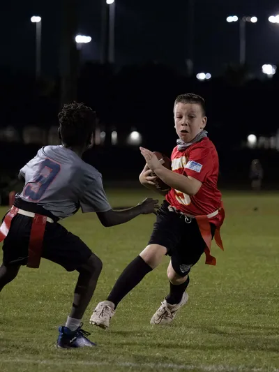 Youth flag football player in red jersey carrying the ball while evading a defender under stadium lights at night