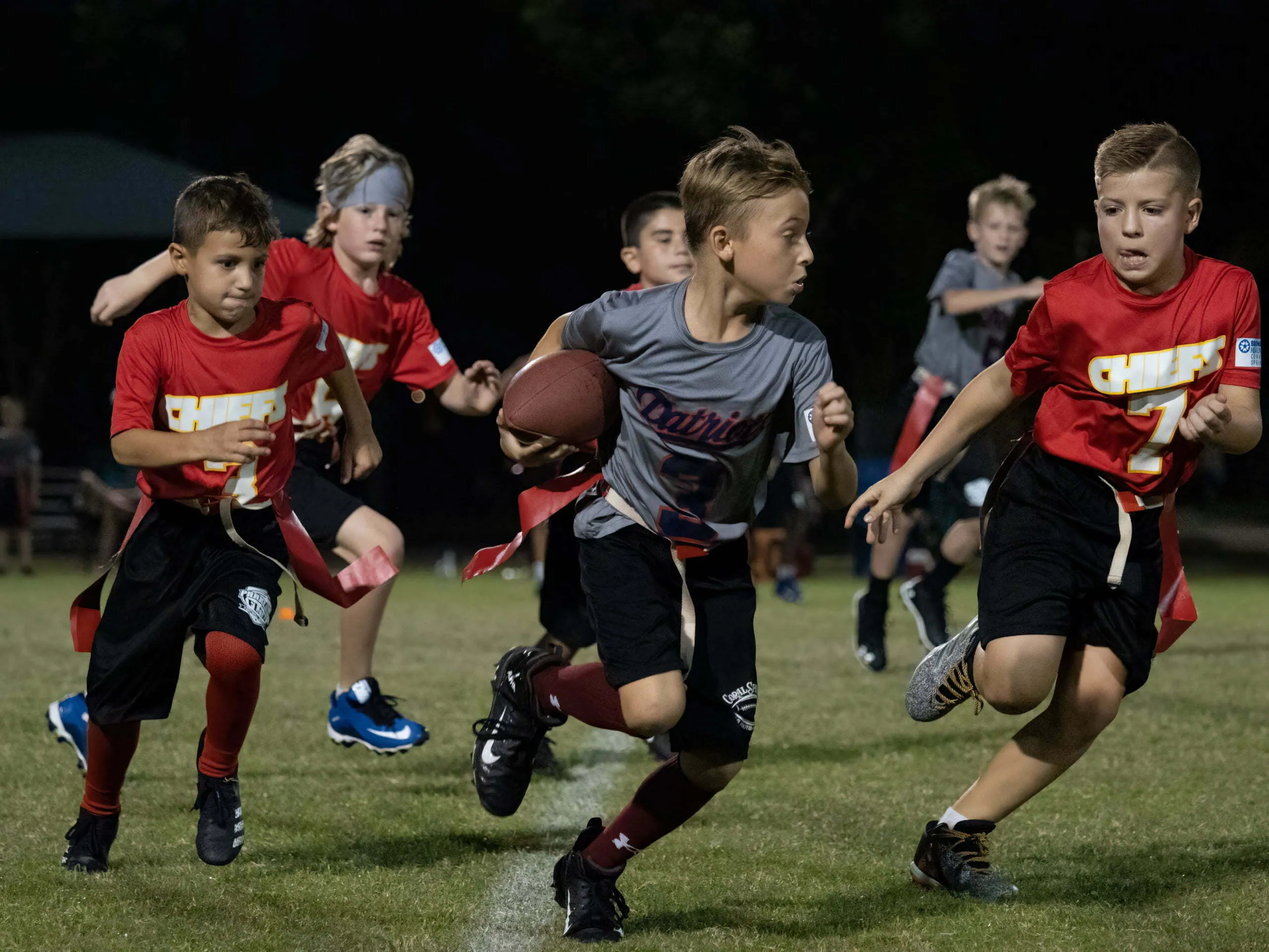 Six youth flag football players in red and gray jerseys sprinting across the field during a nighttime game