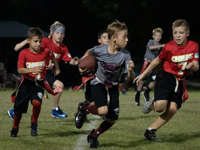 Six youth flag football players in red and gray jerseys sprinting across the field during a nighttime game