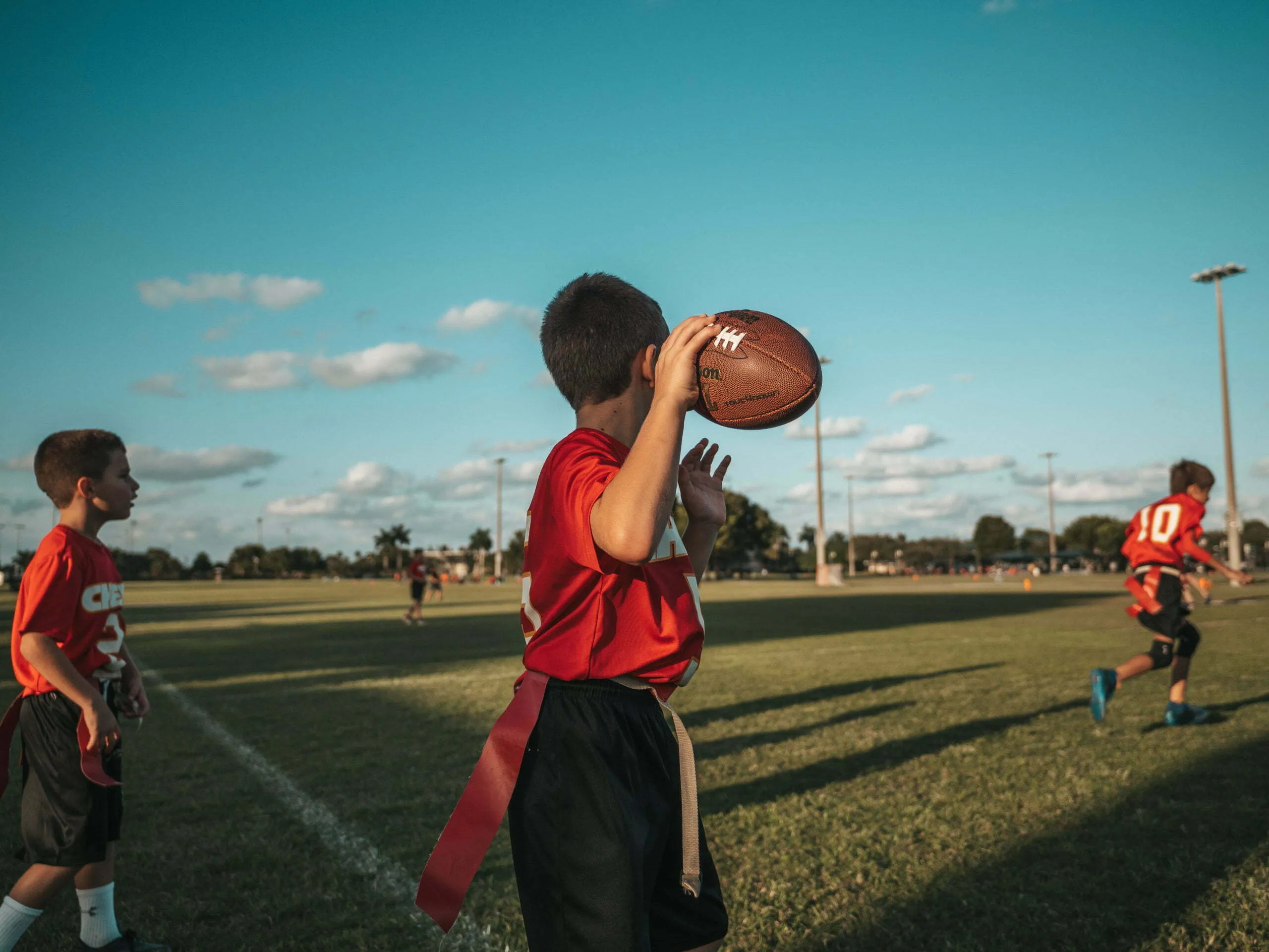 Youth flag football player in red jersey winding up to throw the ball on a green field under a blue sky with scattered clouds