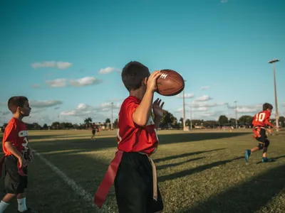 Youth flag football player in red jersey winding up to throw the ball on a green field under a blue sky with scattered clouds