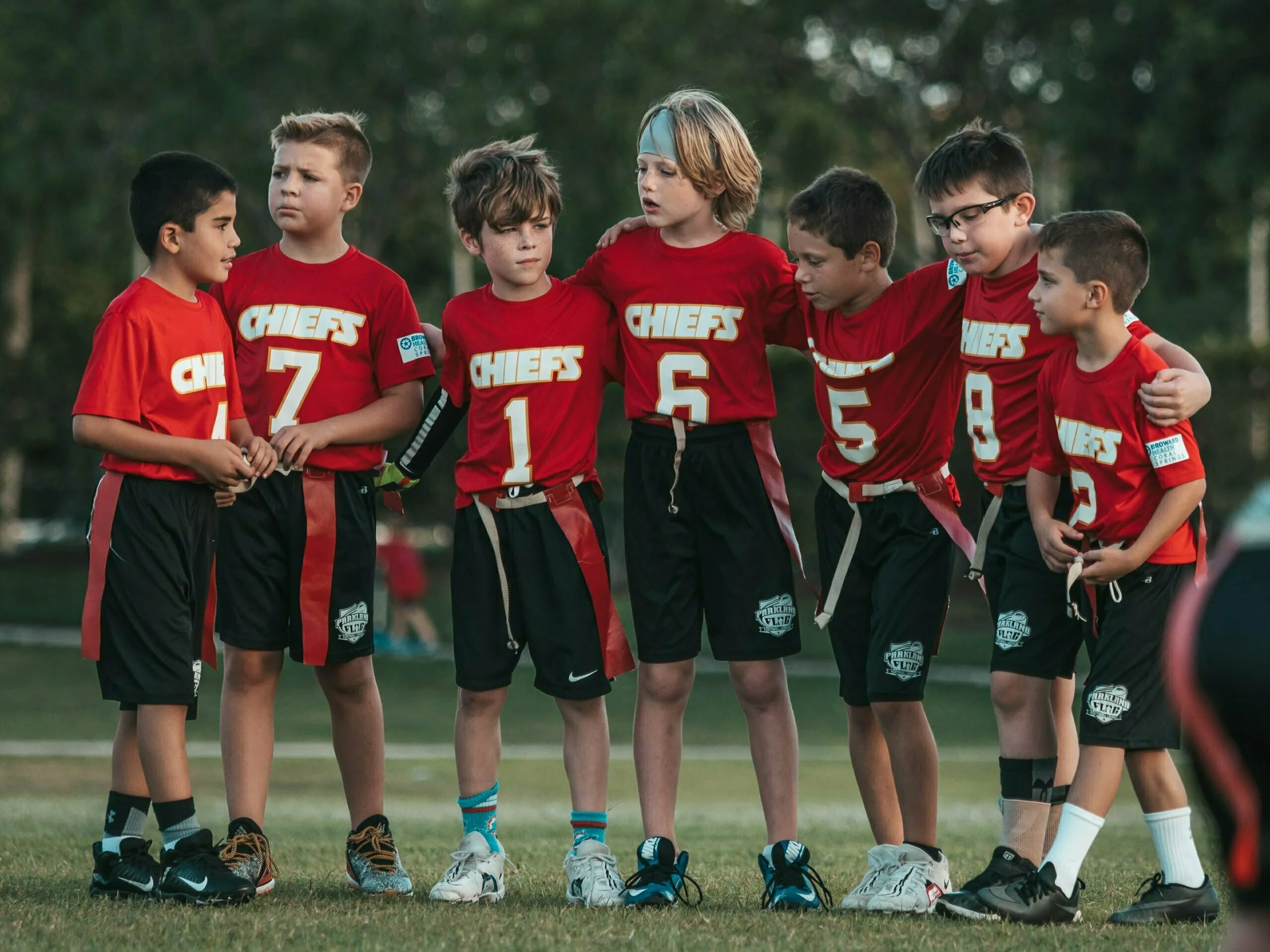 Seven young boys in red Chiefs jerseys huddle together on a grass field during a flag football game at sunset