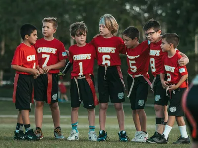 Seven young boys in red Chiefs jerseys huddle together on a grass field during a flag football game at sunset