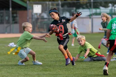Youth flag football player in a Bengals jersey dodging defenders on a green field at golden hour