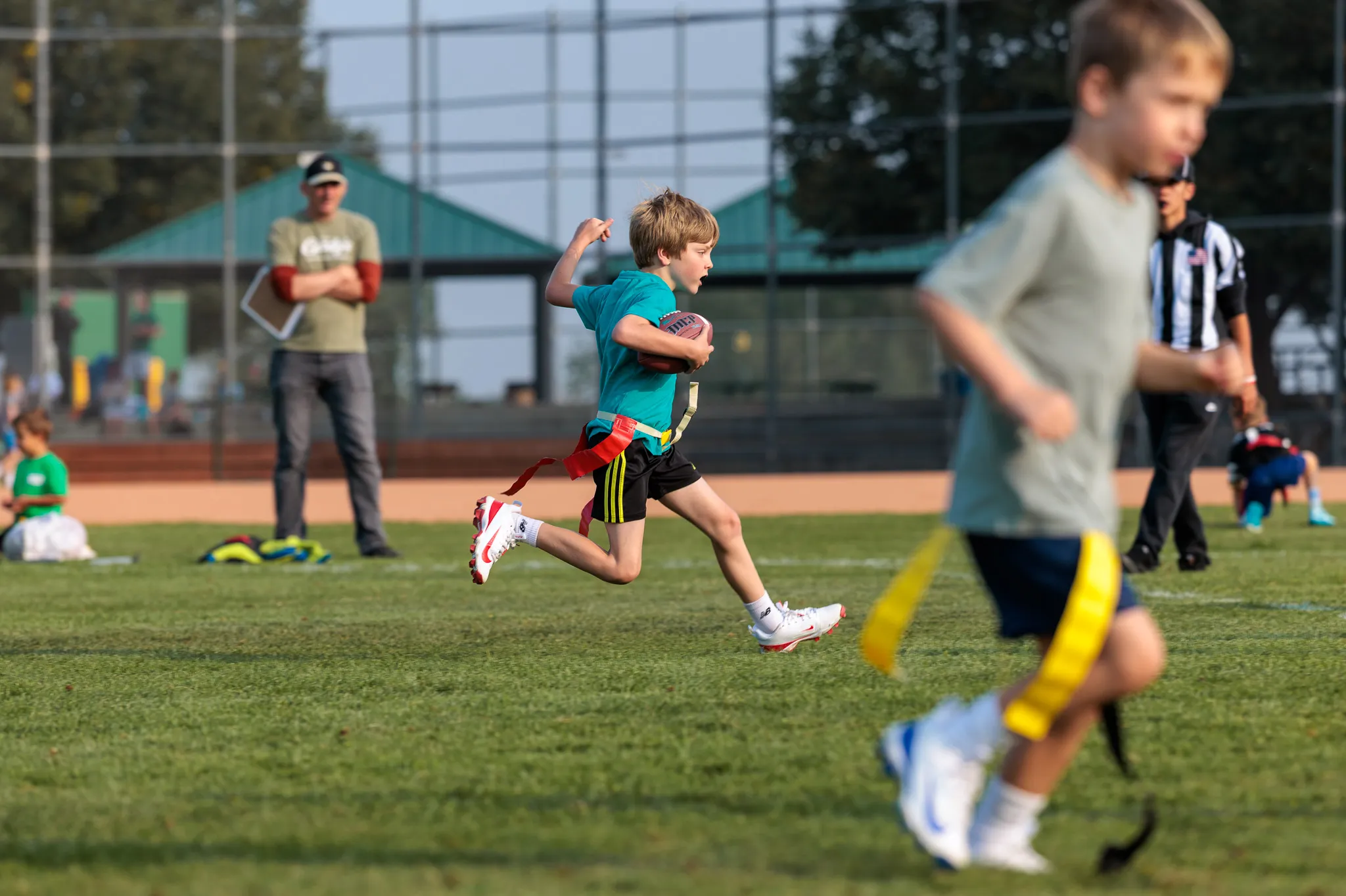Young boy in teal shirt sprinting with football during flag football game on grass field with spectators