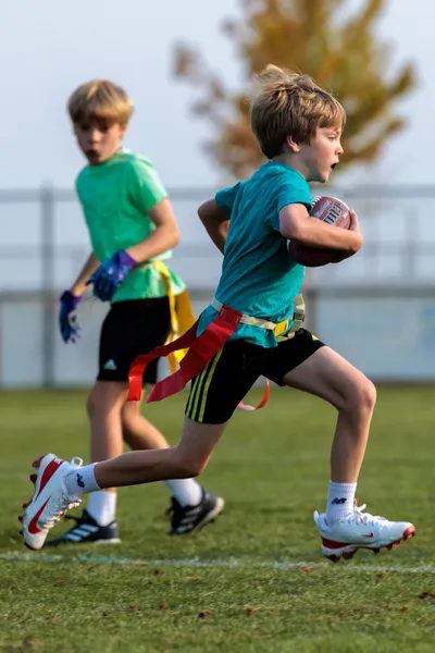 Boy in teal jersey carrying football with red flag belt during youth flag football, teammate trailing behind