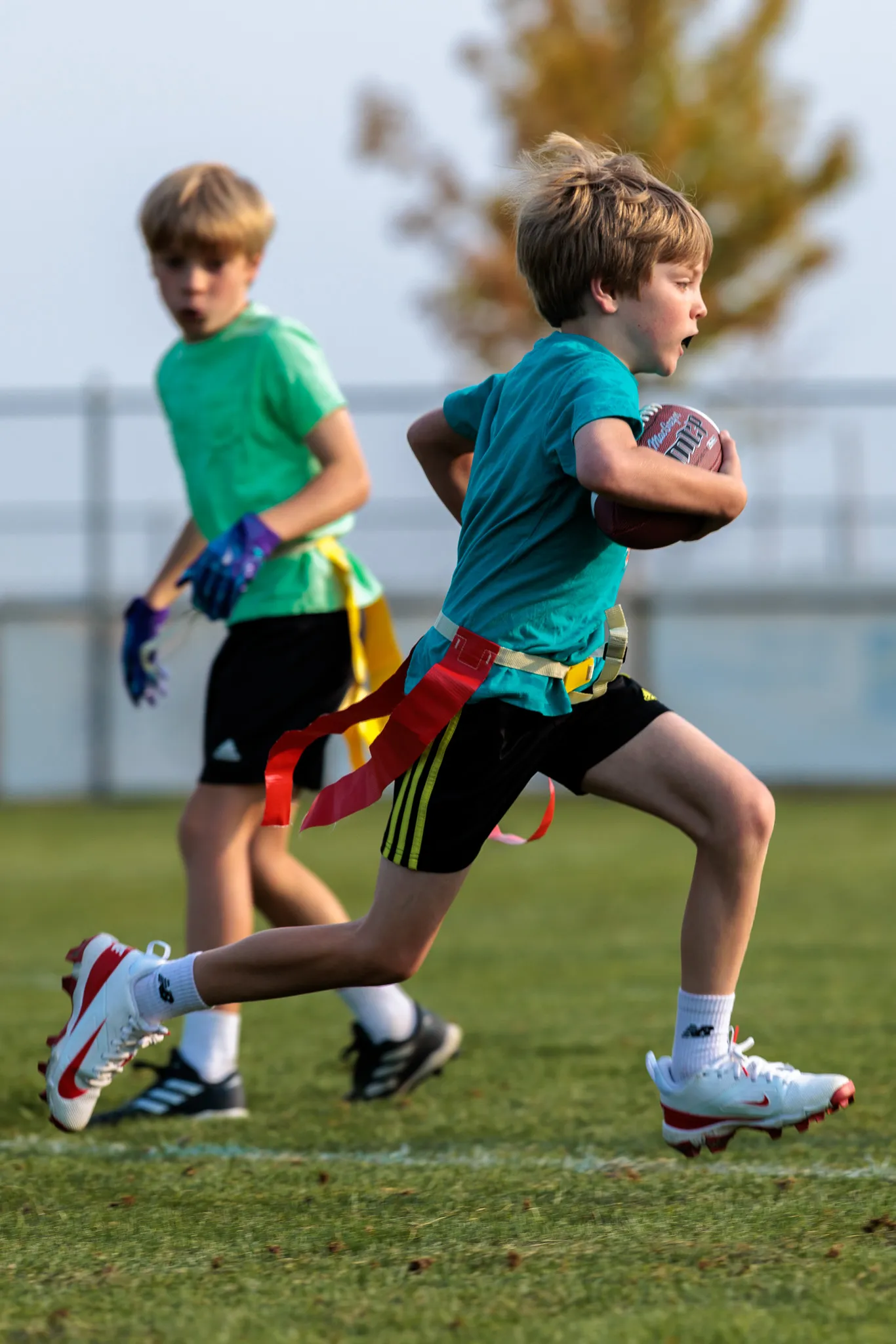 Boy in teal jersey carrying football with red flag belt during youth flag football, teammate trailing behind