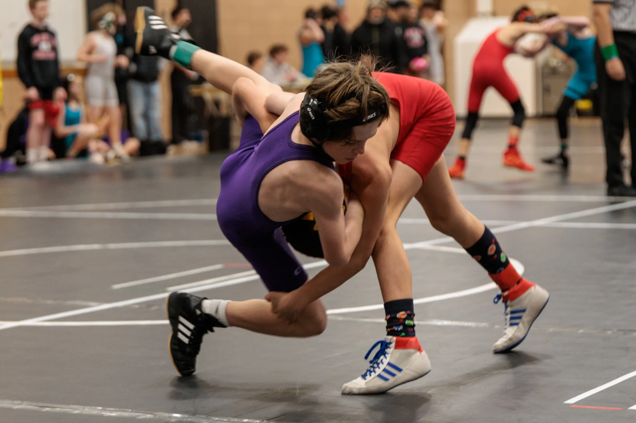 Two youth wrestlers grappling on a gray mat, one in purple singlet and one in red, during an indoor tournament