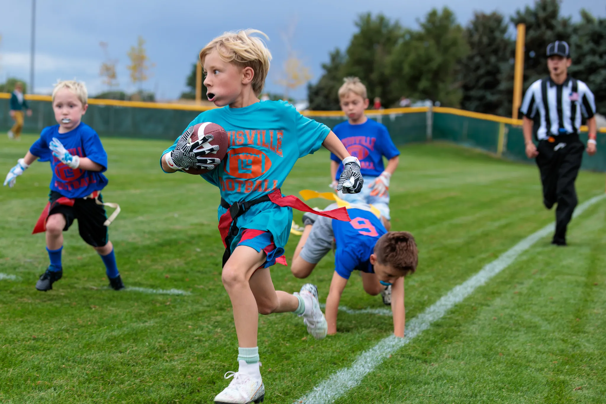 Young boy in teal jersey sprints with football past diving defenders during youth flag football game on green turf field