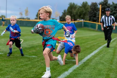 Young boy in teal jersey sprints with football past diving defenders during youth flag football game on green turf field
