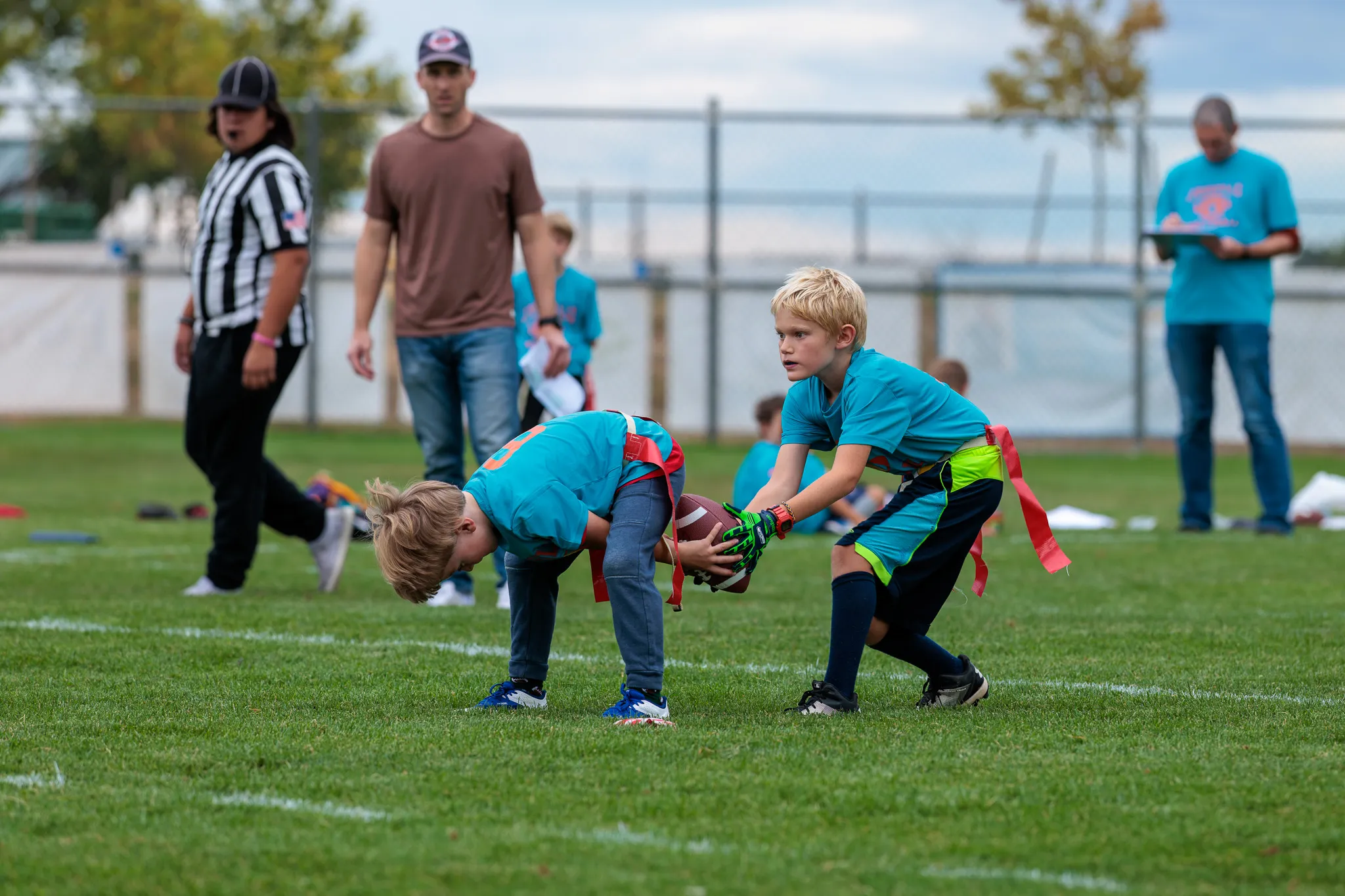 Two boys in teal jerseys execute a handoff during youth flag football with referee and spectators on the sideline