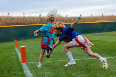 Two youth flag football players race toward the end zone pylon, one in teal and one in blue diving to pull the flag