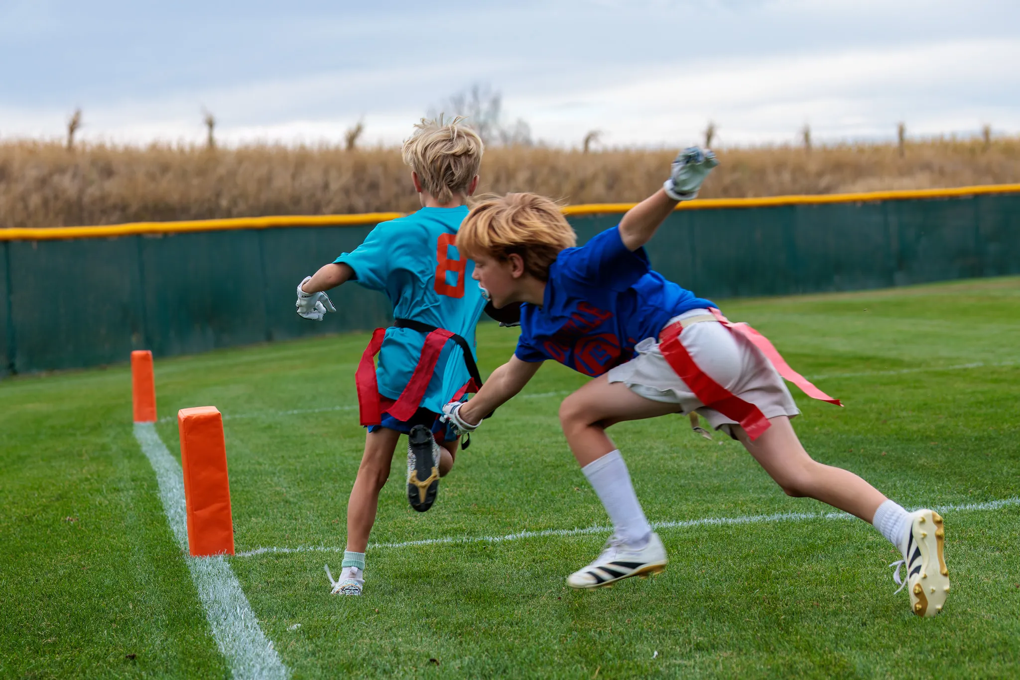 Two youth flag football players race toward the end zone pylon, one in teal and one in blue diving to pull the flag