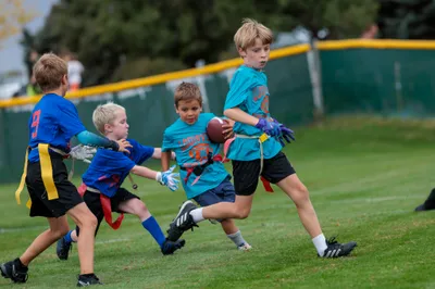 Four young boys scramble for the football during a youth flag football game, teal and blue jerseys colliding on green turf