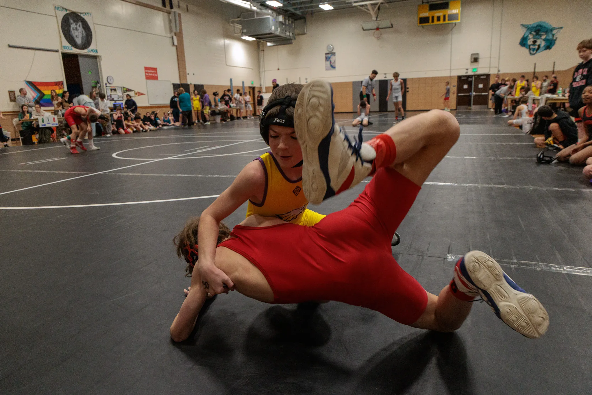 Youth wrestler in gold singlet and headgear takes down opponent in red singlet on a dark gray mat at an indoor tournament
