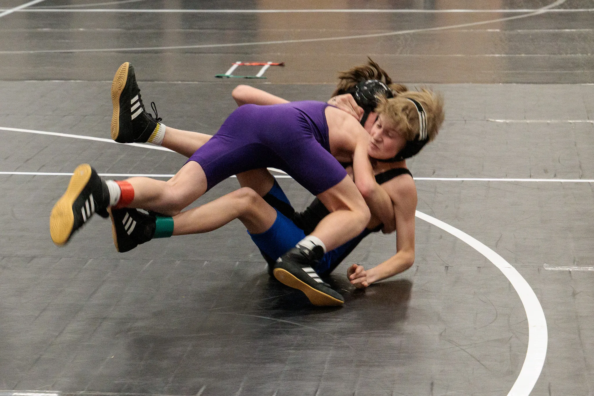 Two youth wrestlers grapple on a dark mat, one in purple singlet controlling an opponent in black from above