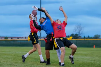 Three teens leap for a contested football catch at dusk during a flag football game, blue and red jerseys against a twilight sky