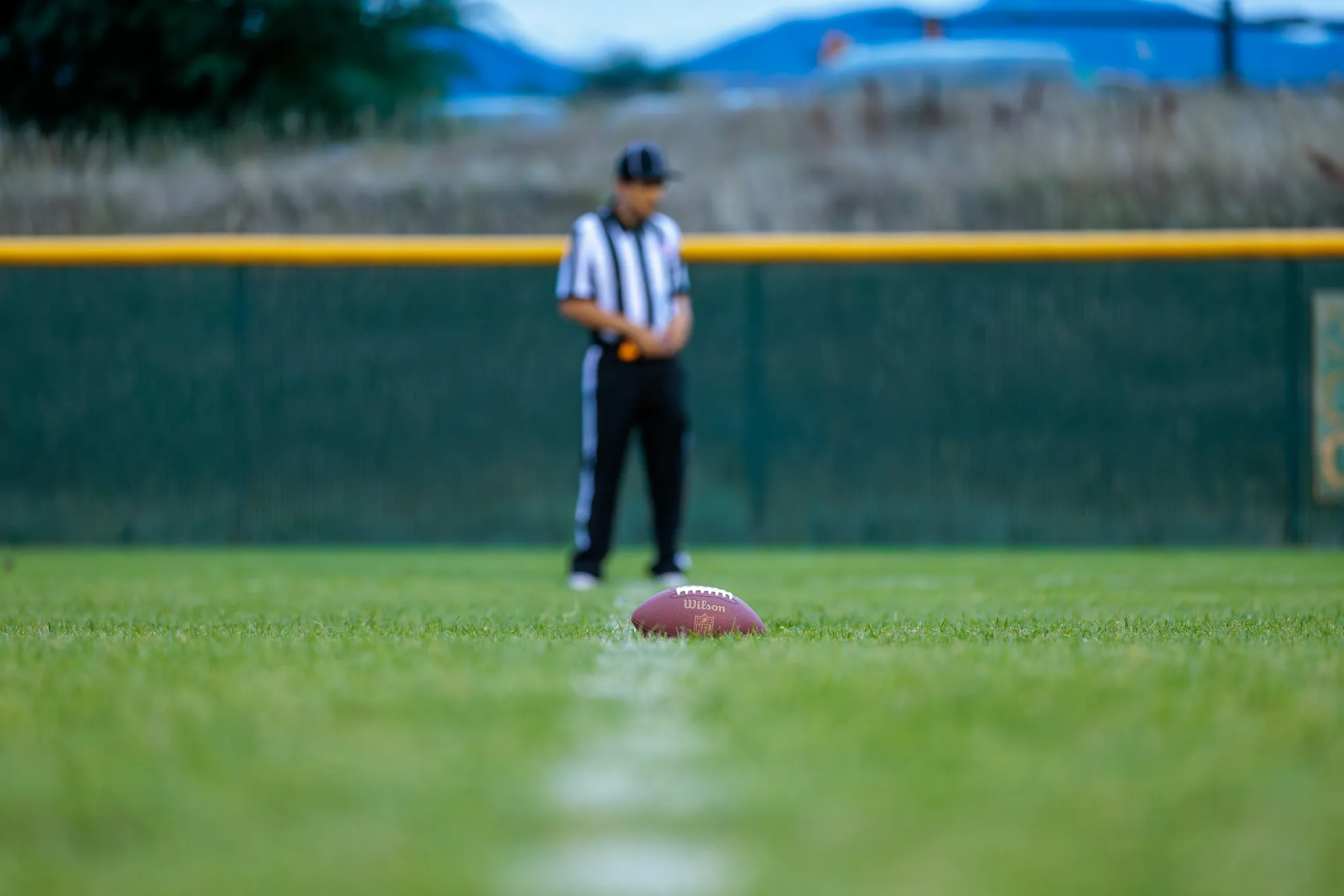 Football resting on a white yard line with a blurred referee standing in the background on a green turf field at dusk