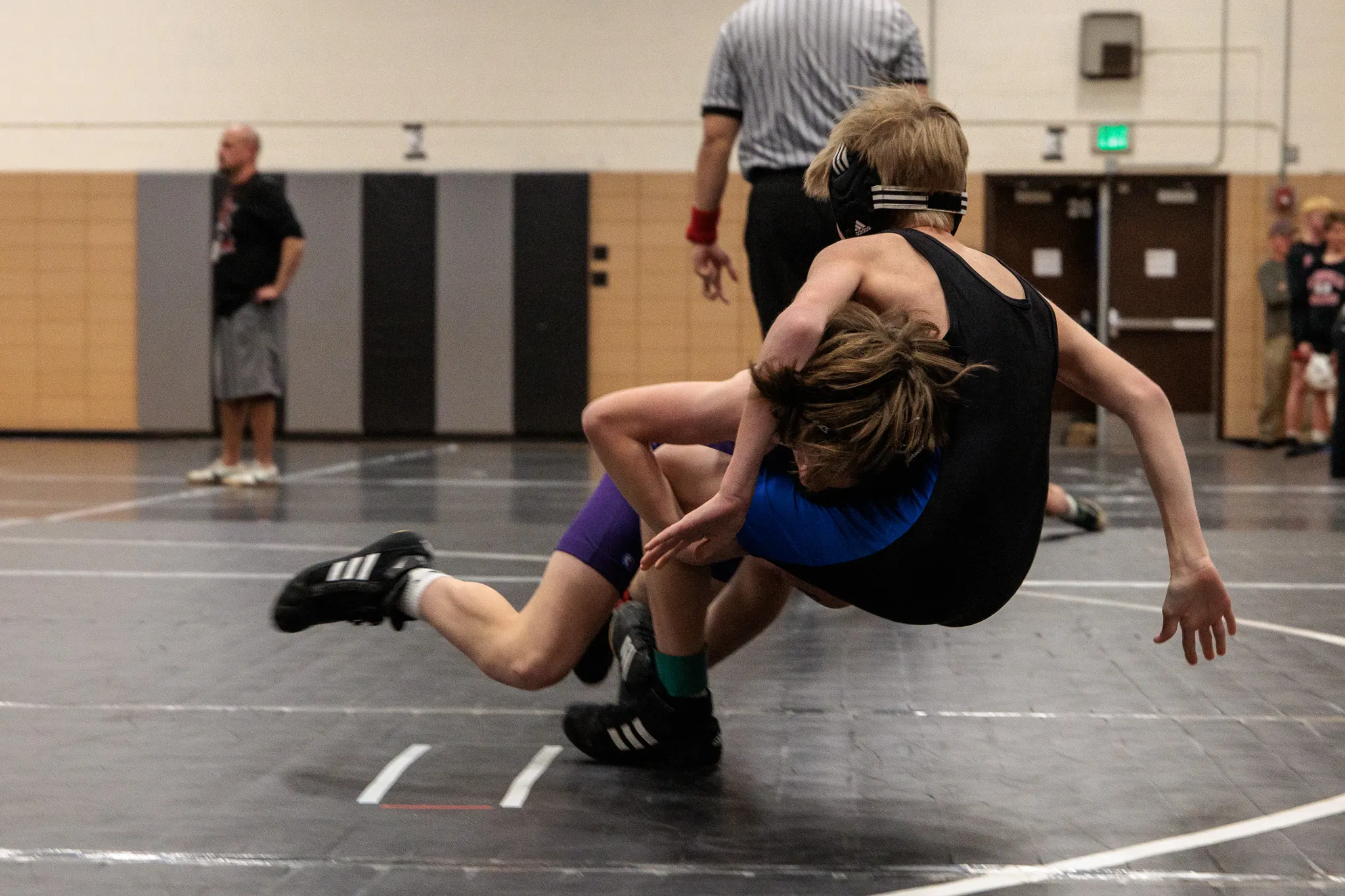 Two youth wrestlers mid-takedown on a black mat, one in purple singlet driving opponent in blue to the ground