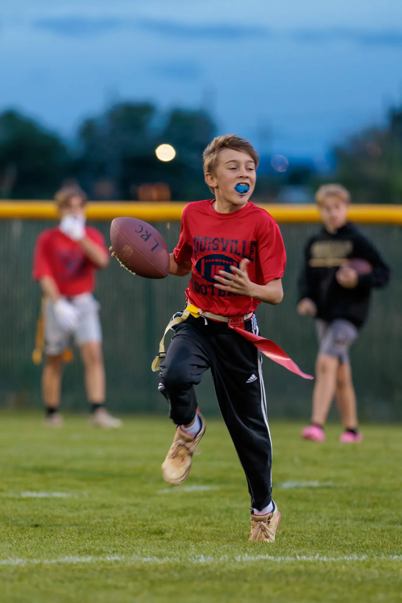 Boy in red Louisville shirt running with football and blue mouthguard during youth flag football game at dusk