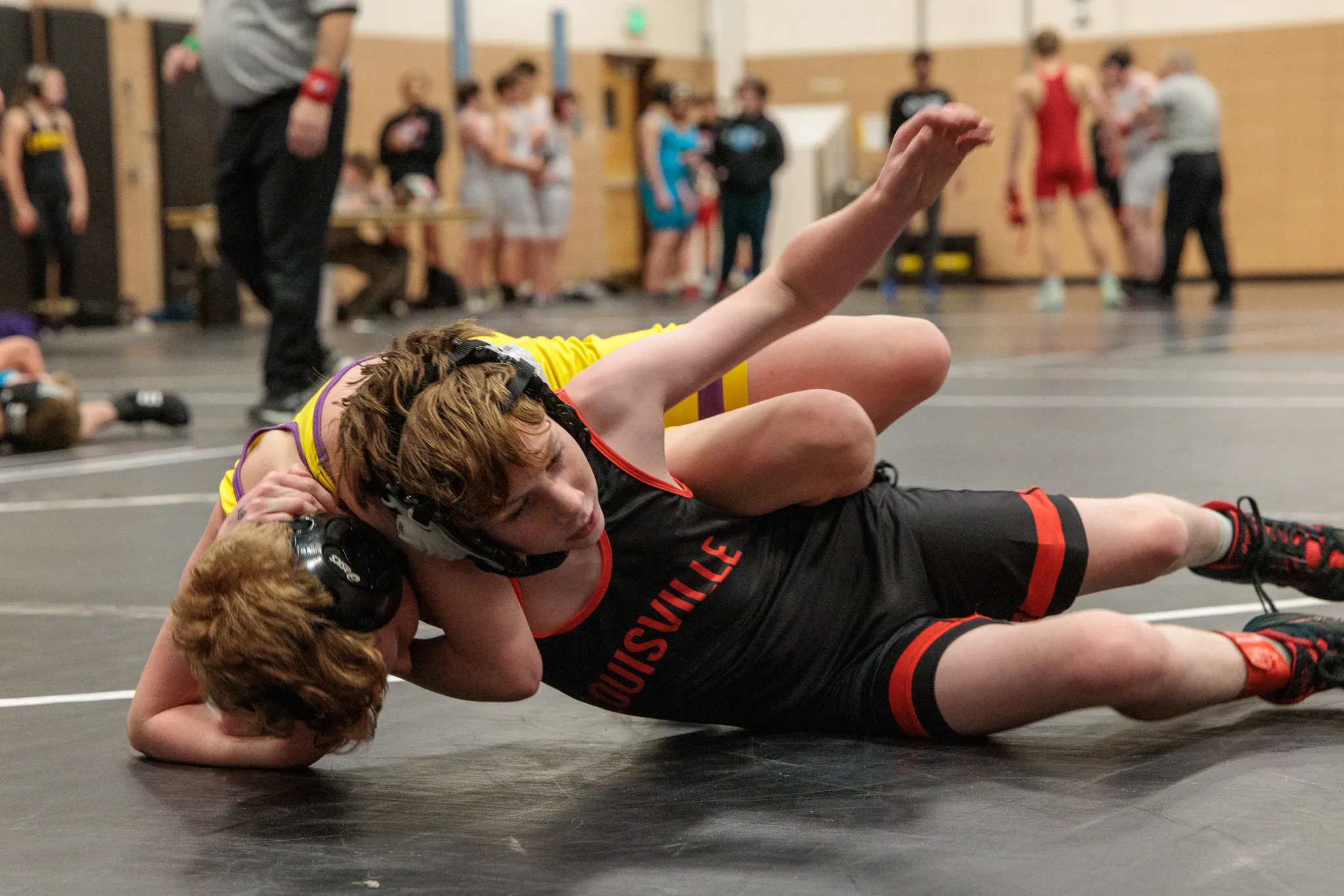 Two youth wrestlers on the mat in a pin attempt, wrestler in black Louisville singlet controlling opponent with headgear