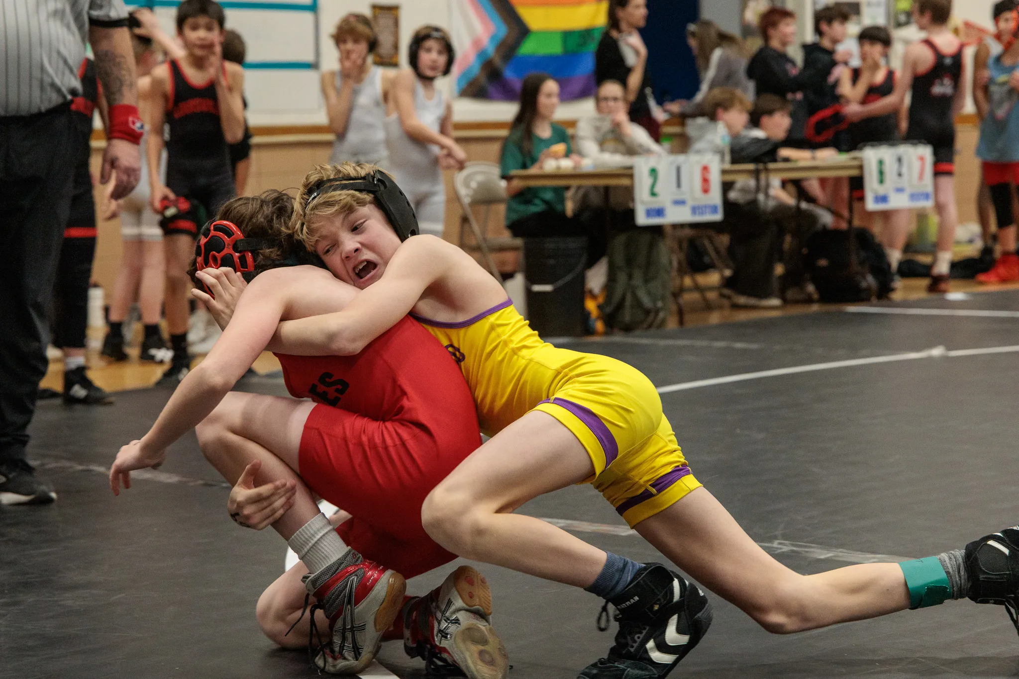 Two youth wrestlers locked in a scramble on the mat, one in red and one in yellow singlet with crowd watching closely