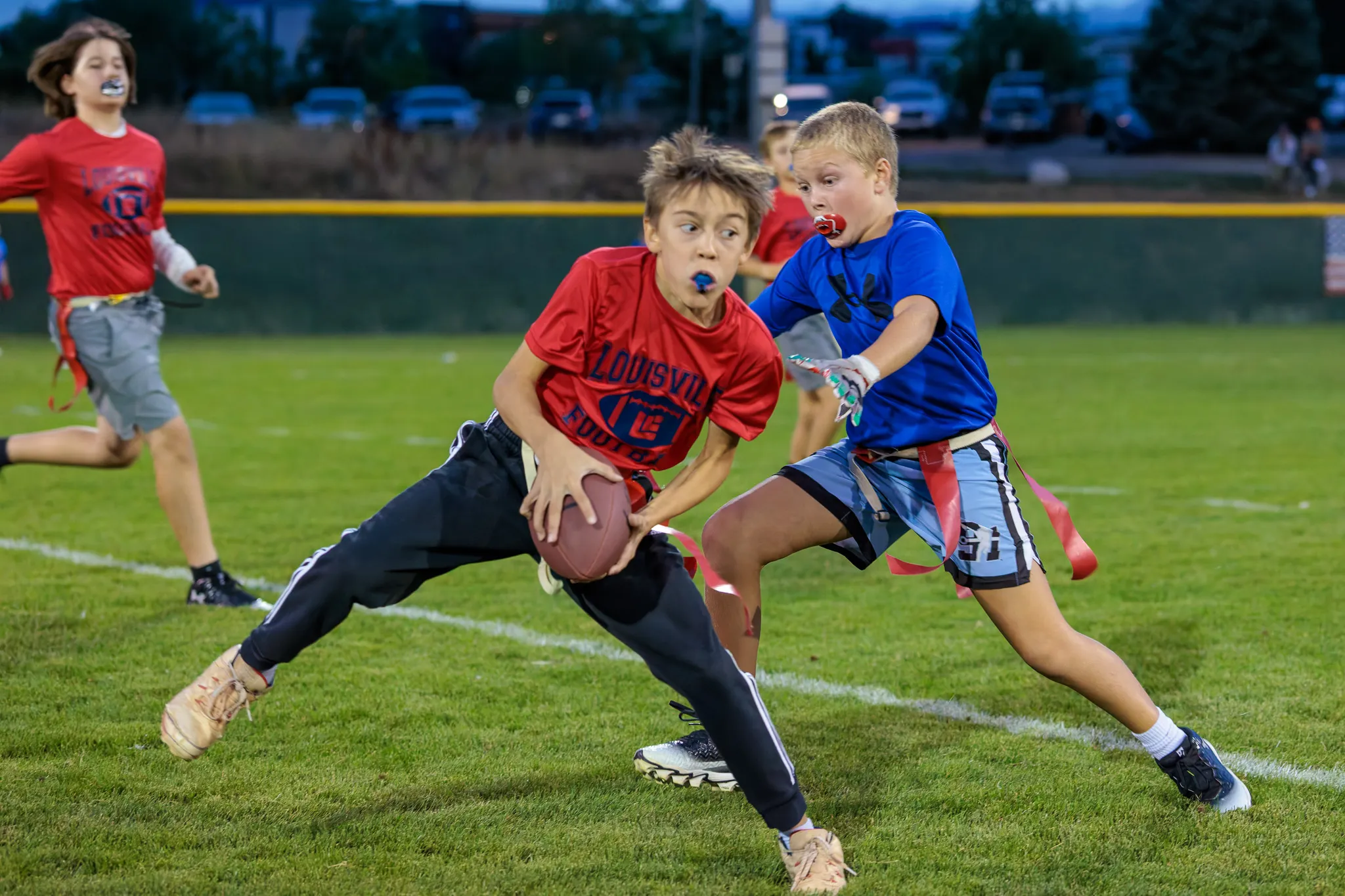 Youth flag football player in red dodging a defender in blue while carrying the ball on a grass field at dusk