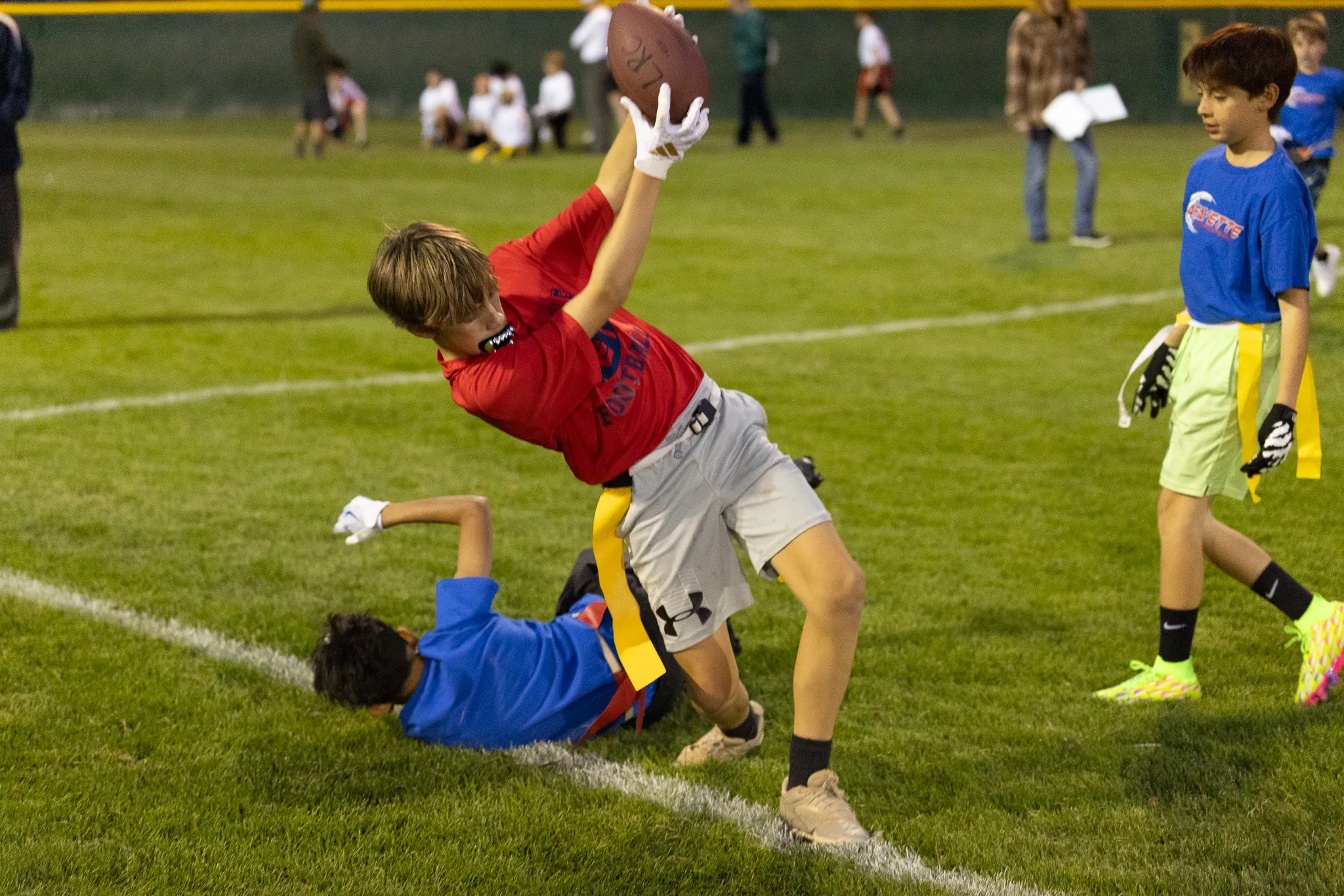 Youth flag football player in red jersey leaping to catch a football near the sideline under stadium lights