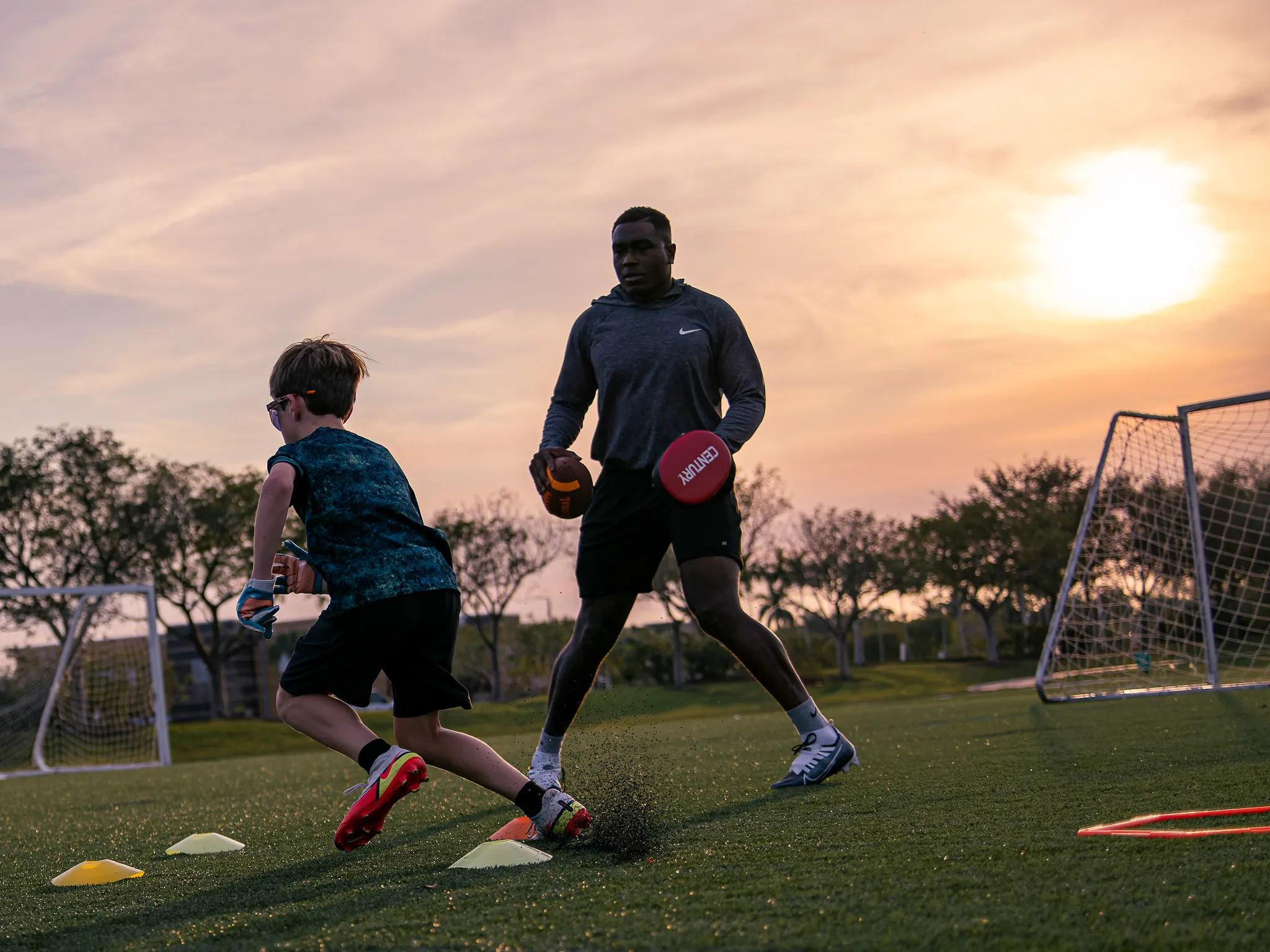 Youth football training session at sunset with coach holding ball and boy running agility drill around cones on turf field