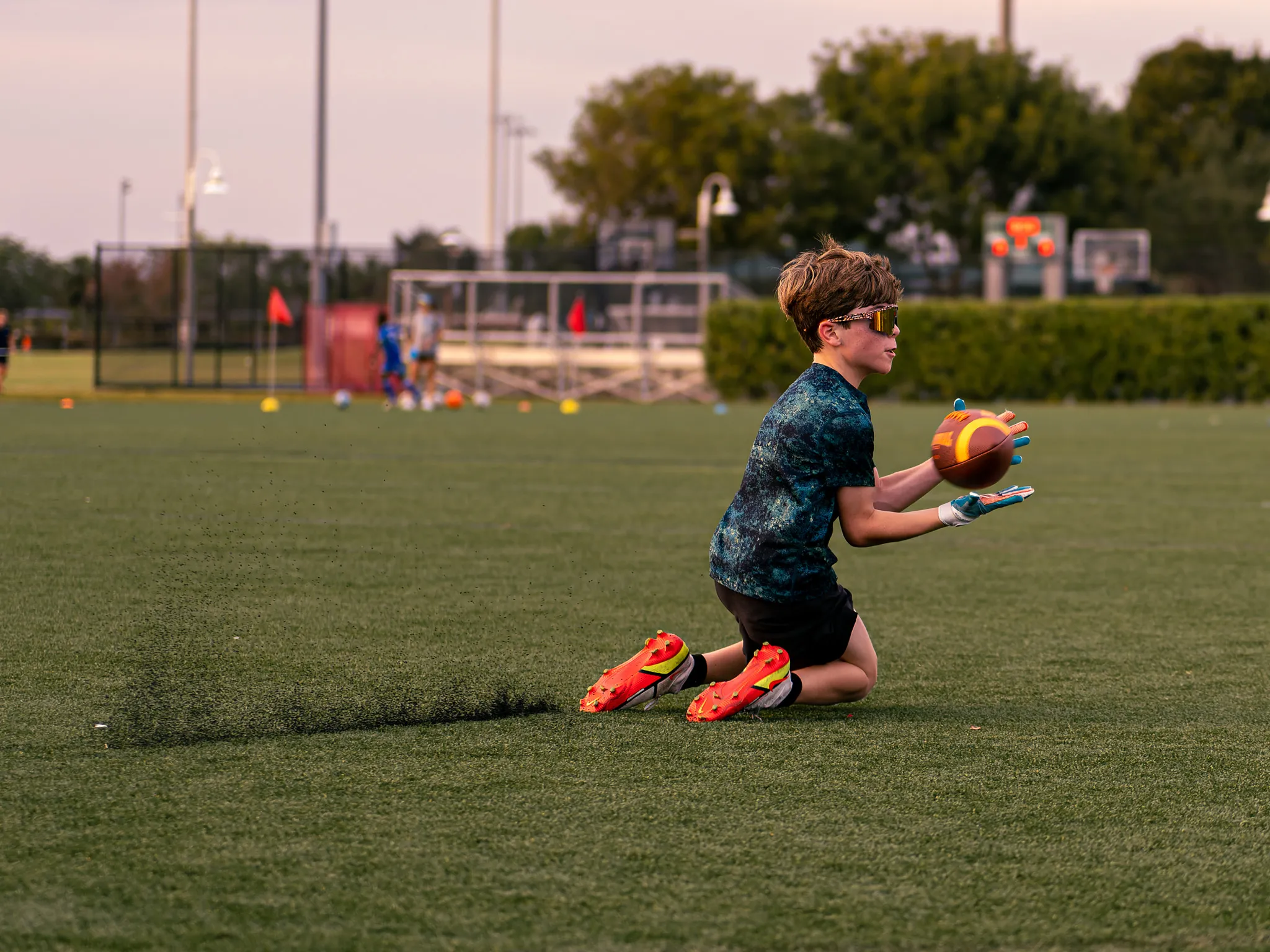 Boy kneeling on turf field catching a football during training session at dusk wearing yellow sunglasses and bright cleats