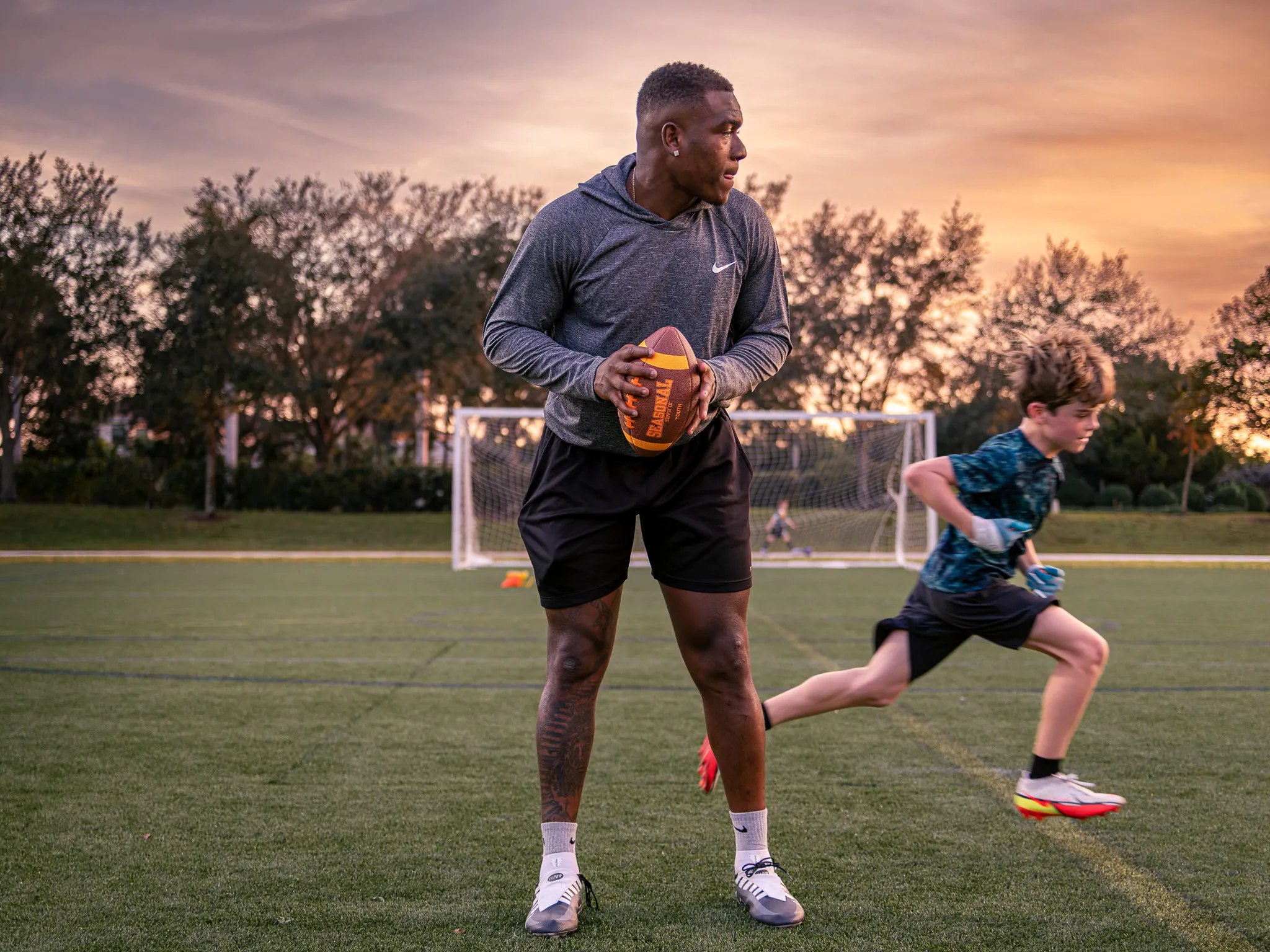 Athletic coach standing on turf field holding football at sunset while young player sprints behind him near goal posts