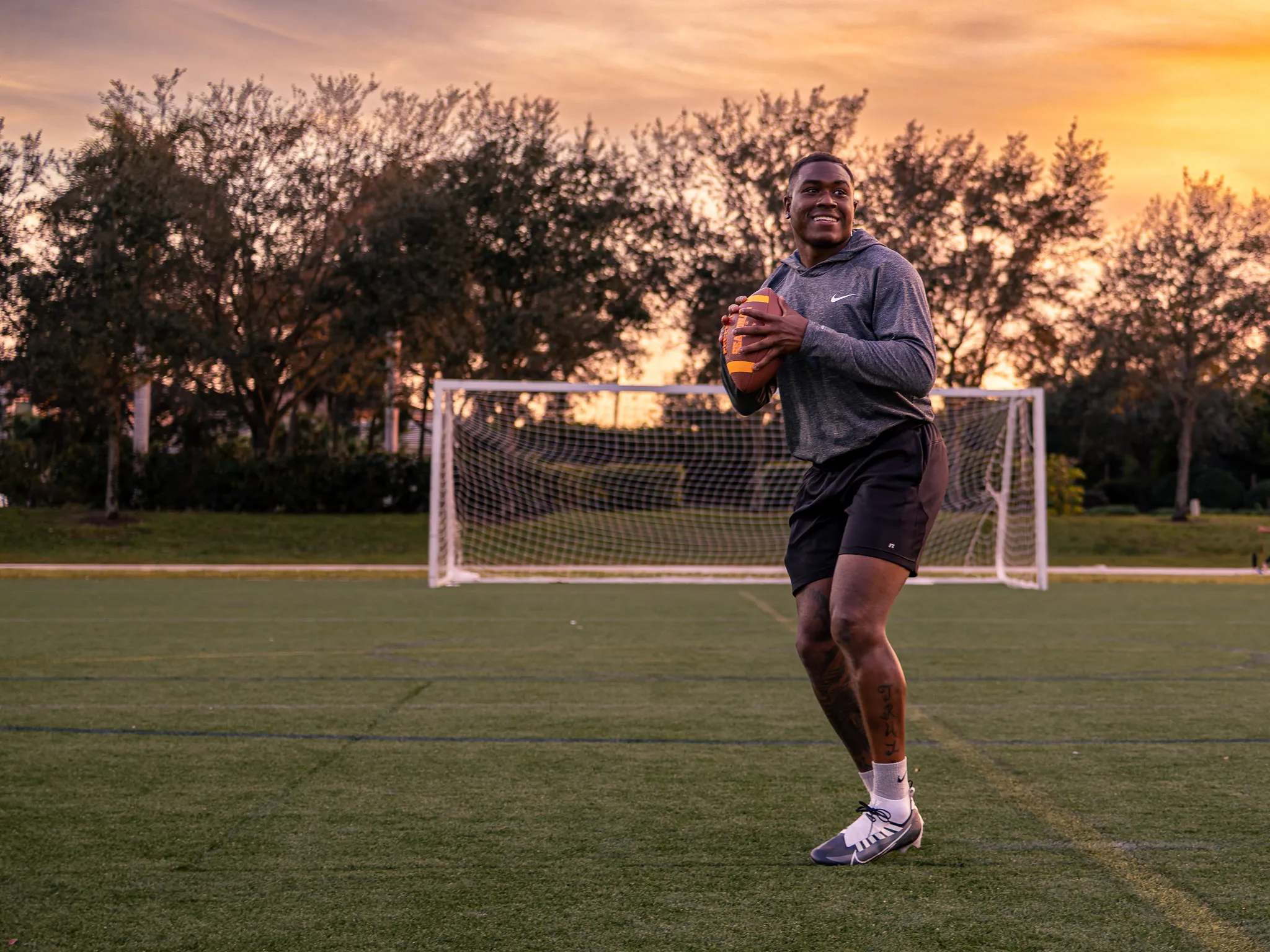Smiling athlete in gray hoodie throwing football on turf field with orange sunset sky and trees behind him