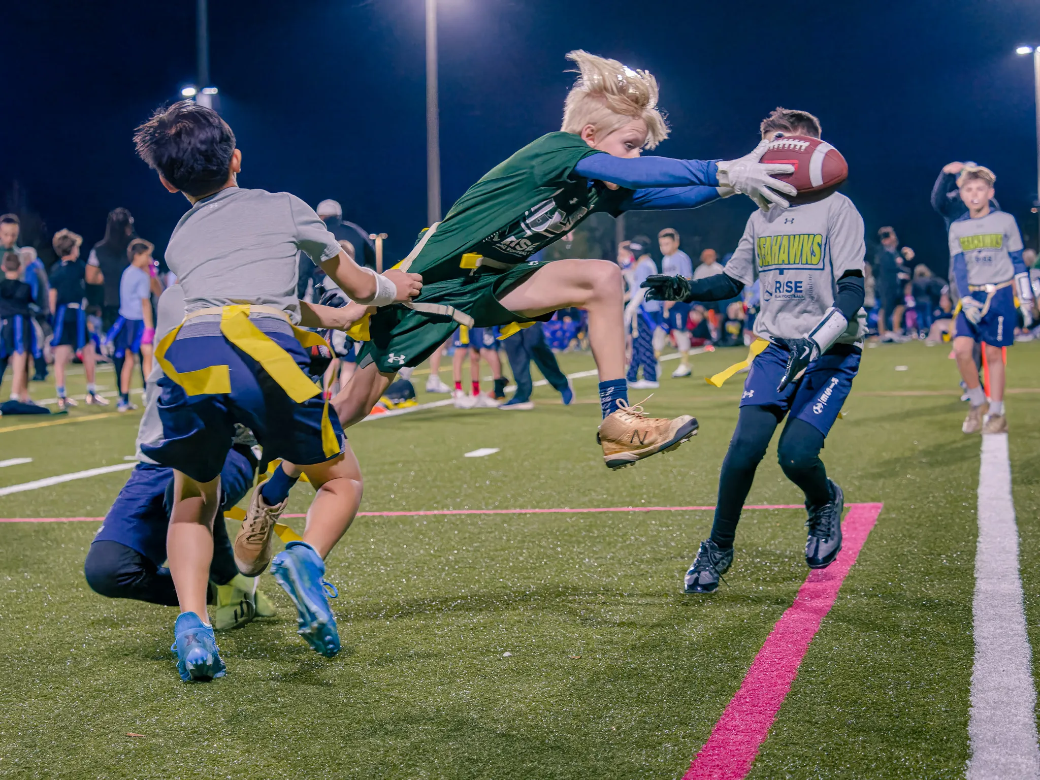 Youth flag football player in green jersey leaping to catch a one-handed pass while defenders in blue and gold reach to tag