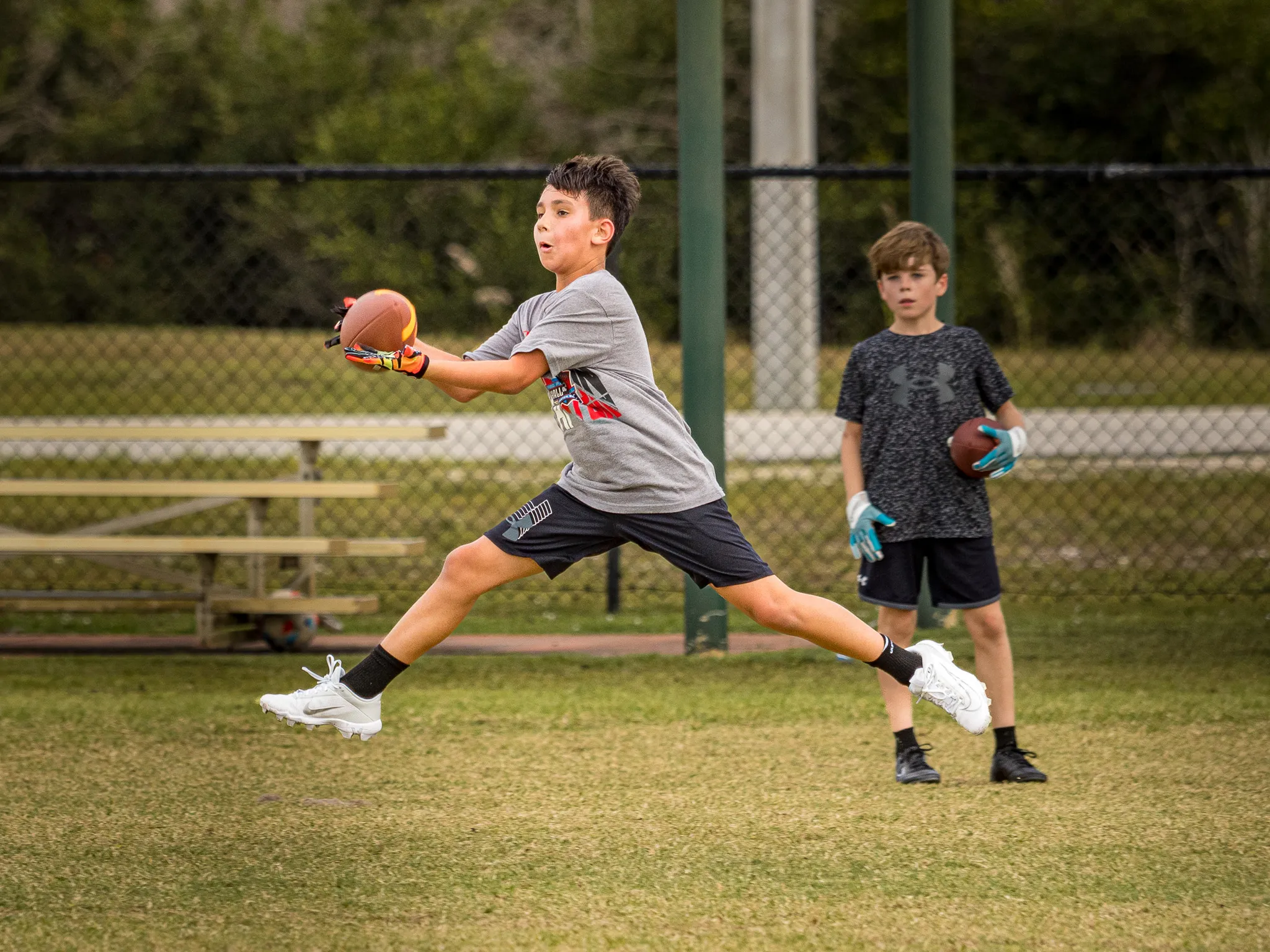 Teen boy lunging to catch a football on a grass field while another player watches with a ball nearby