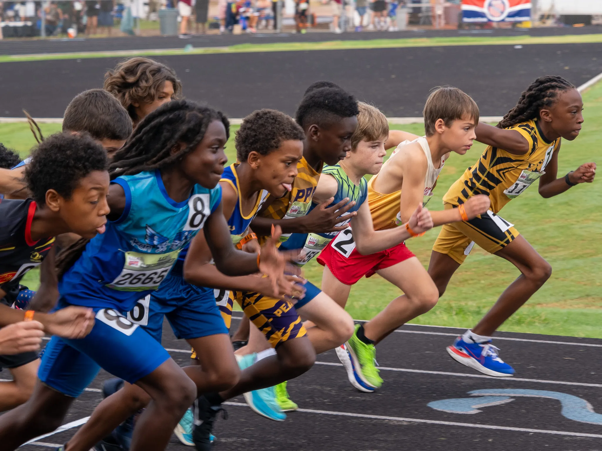 Youth track and field runners exploding off the starting line at a meet, roughly ten kids in colorful jerseys