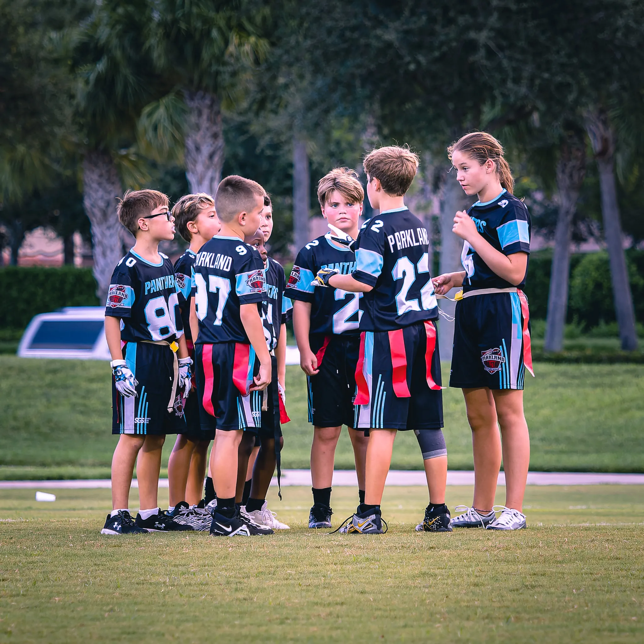 Seven youth flag football players in black Parkland jerseys huddling on a green field at dusk with palm trees behind them