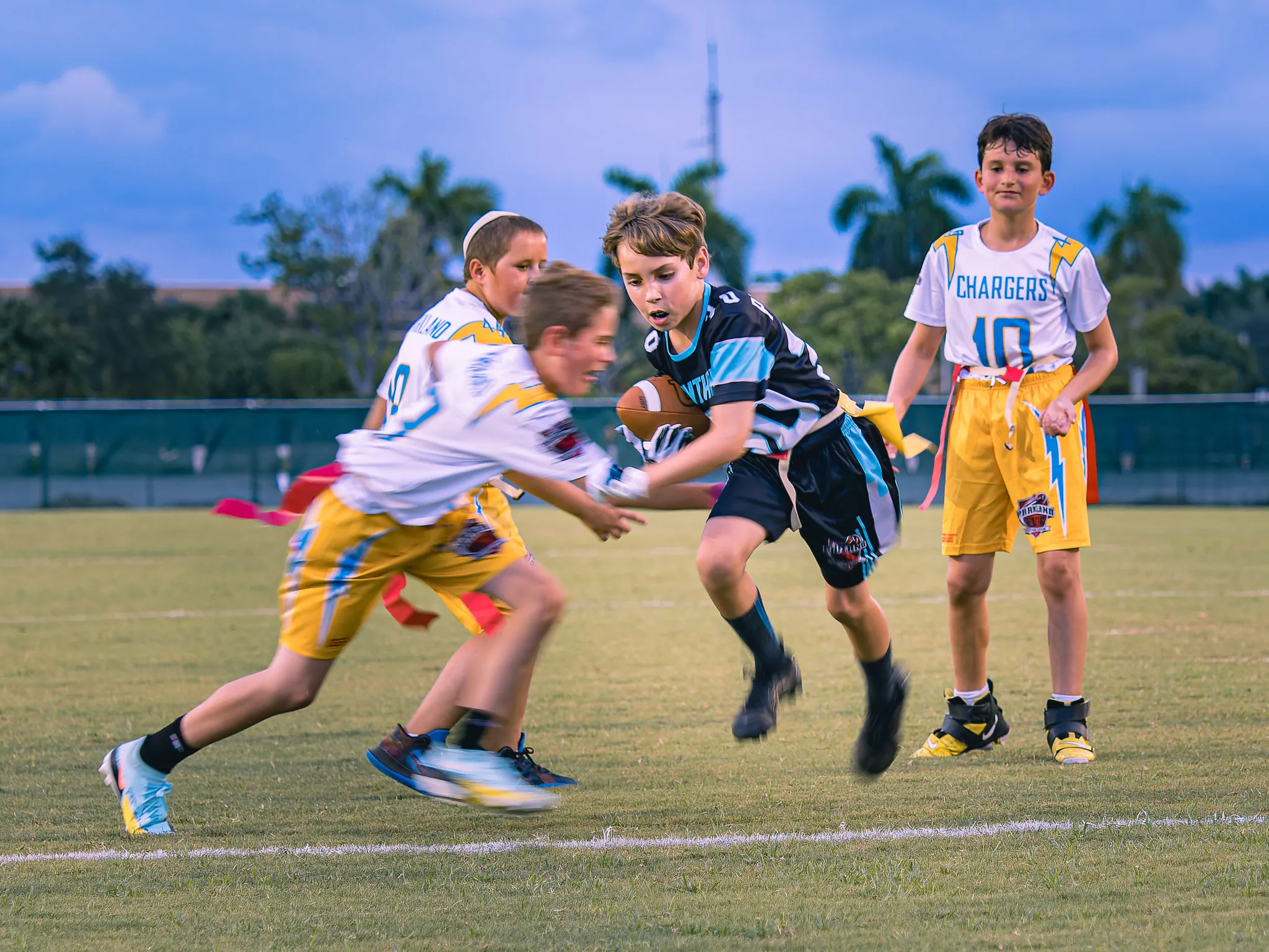 Youth flag football player in dark jersey carrying the ball while evading a defender in a white Chargers jersey on a grass field