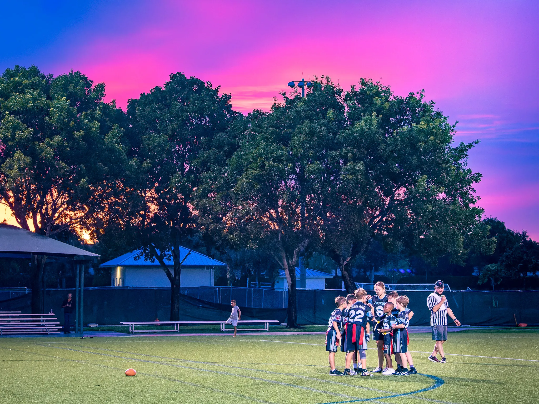 Youth flag football team huddling on a turf field under a vivid pink and purple sunset sky with large trees silhouetted behind them
