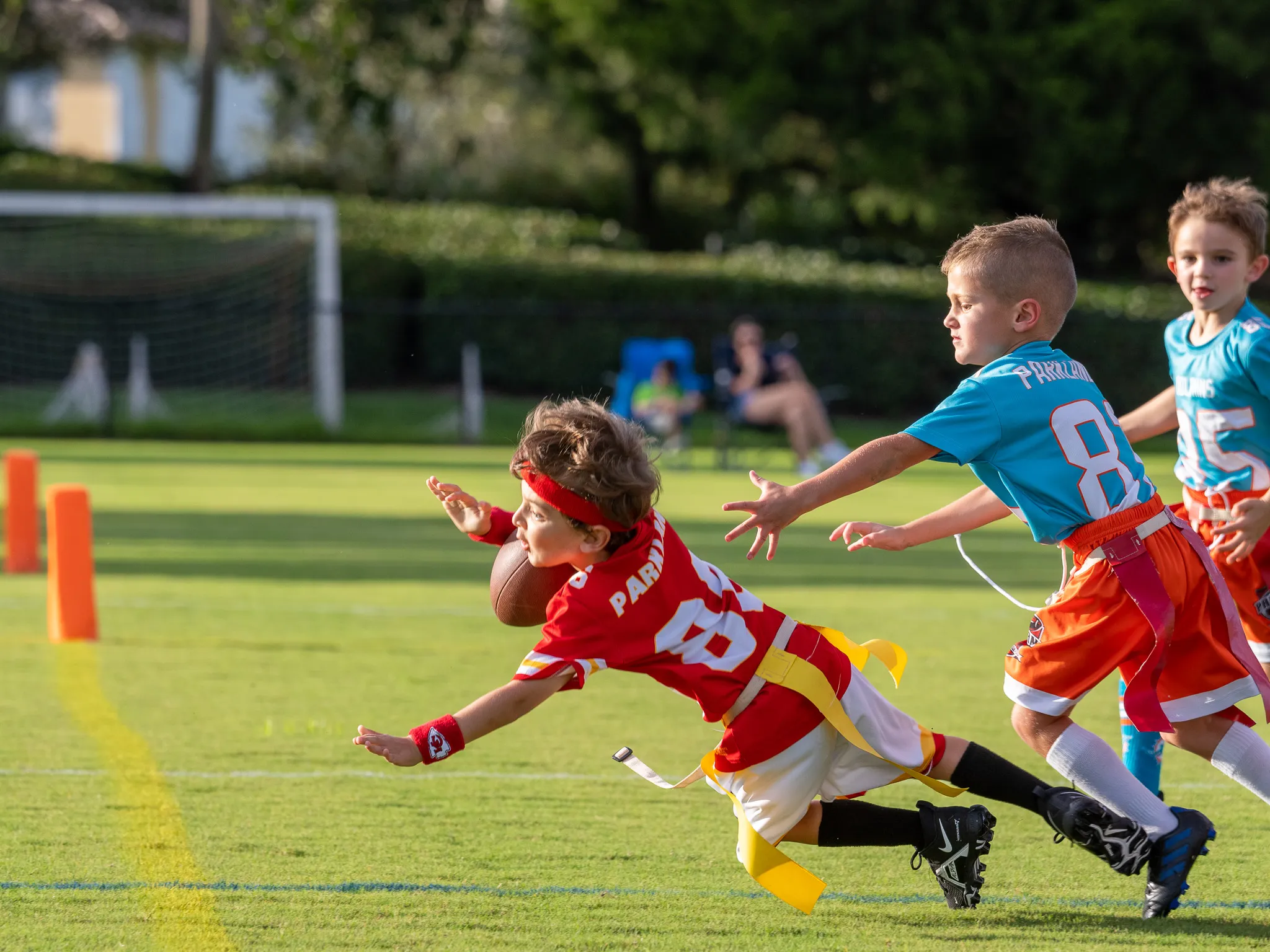 Three young boys playing flag football on a green field, one in red diving for a catch while two in teal pursue