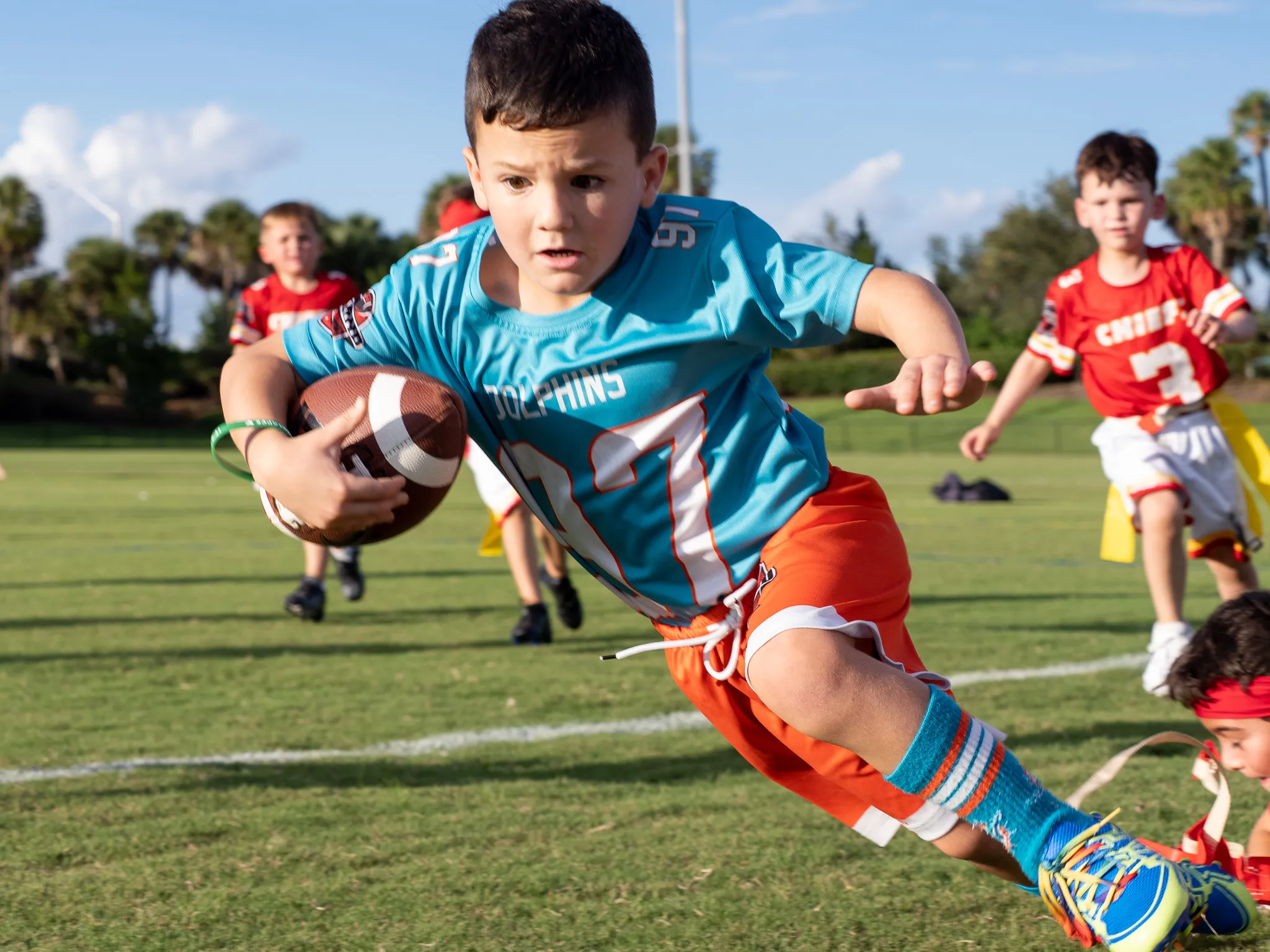 Young boy in teal Dolphins jersey sprinting with football tucked under arm during youth flag football game on turf field