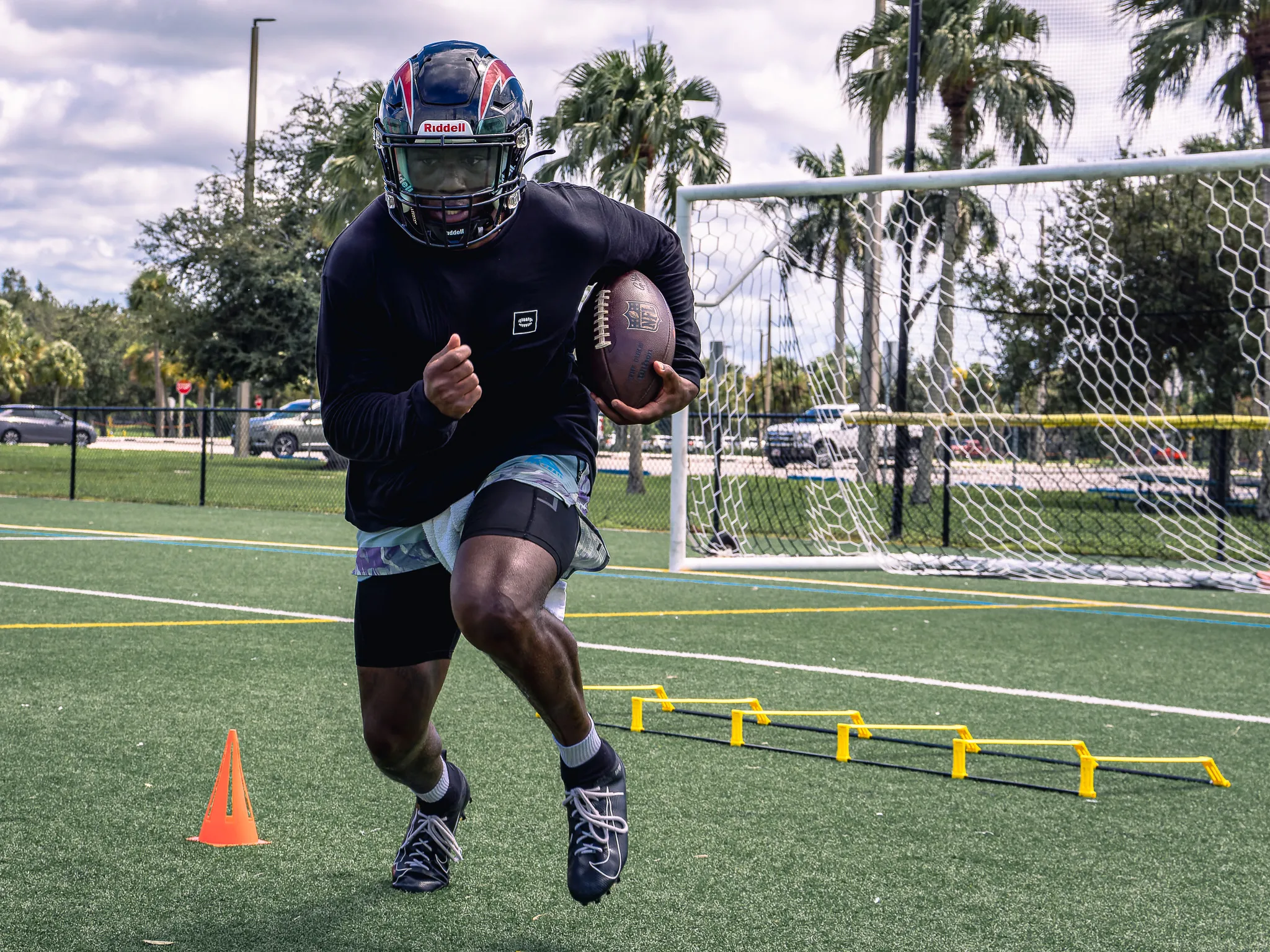 Football player in dark helmet and black training gear running agility drill with ball over yellow hurdles on turf field
