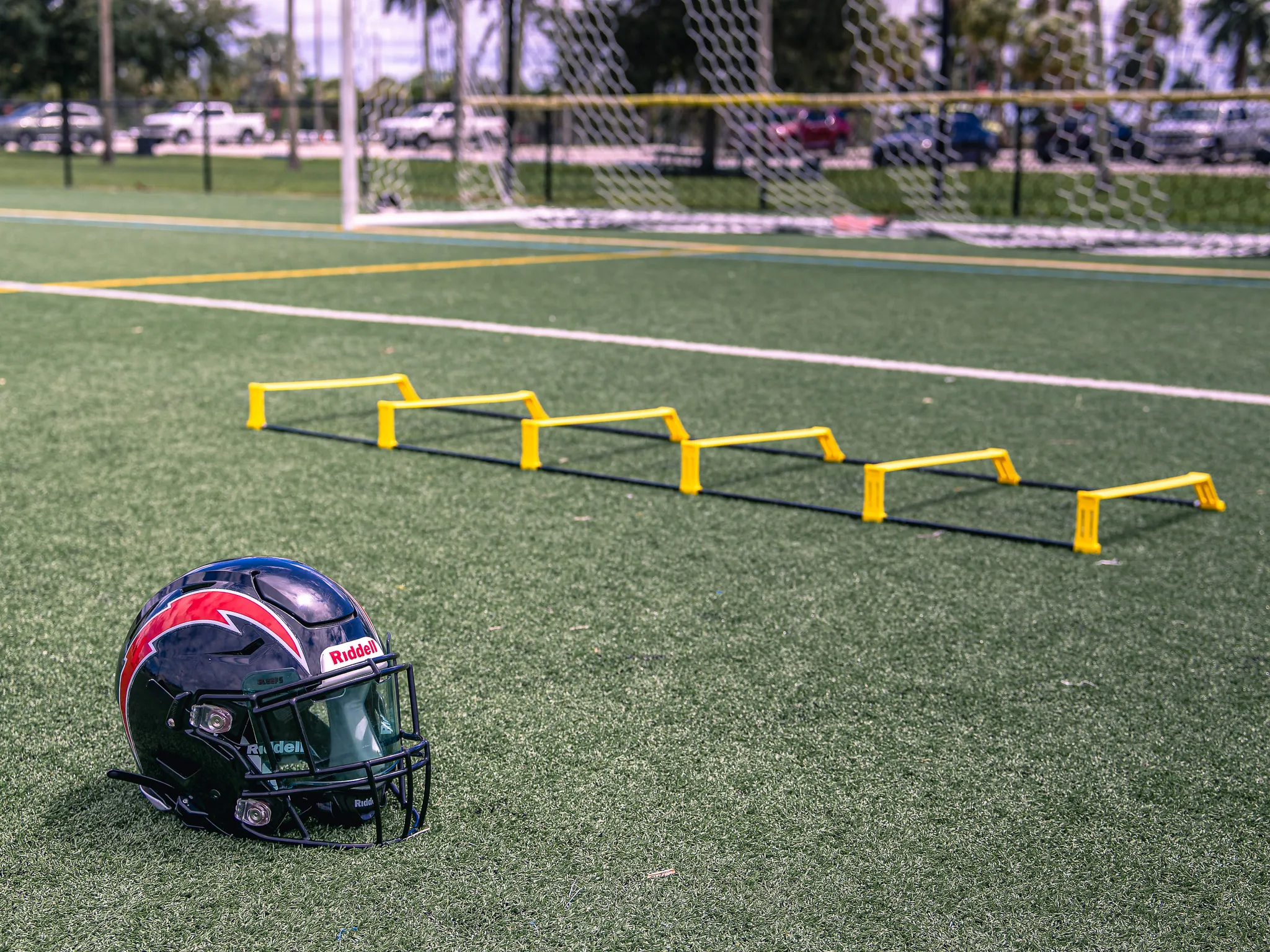 Dark football helmet with red and blue decals resting on green turf beside a row of yellow agility hurdles